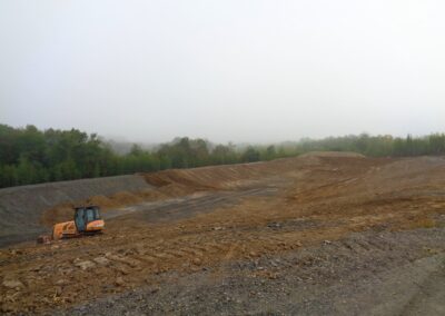 Landscape view of bulldozer spreading piles of topsoil across a rocky basin next to a deciduous forest
