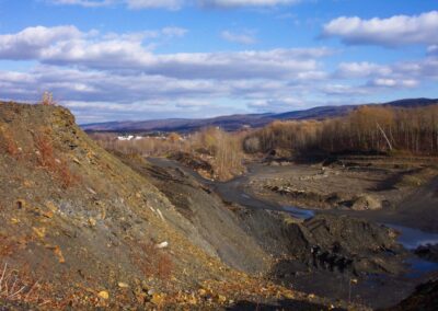Large and steep piles of barren waste rock. Black, silty material covers ground. Trees, residential area, and forested mountains in background