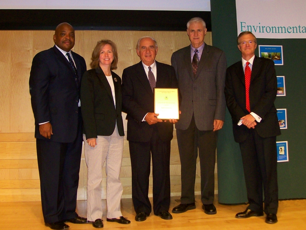 Four men and one woman in suits stand on stage. Man in middle (Mike Dziak) display an honorary plaque