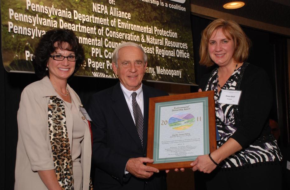 Three adults posed for camera: Woman on right hands framed certificate to man at center, with another woman to his left.