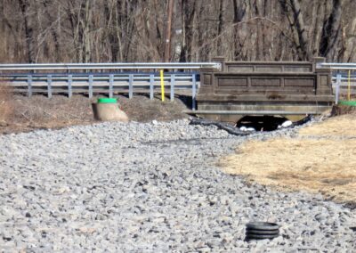 Rock-lined, slightly curved stream channel (without water) headed towards small concrete bridge