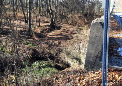 Wall of small concrete bridge on right, looking down bank to overgrown stream with sections without water