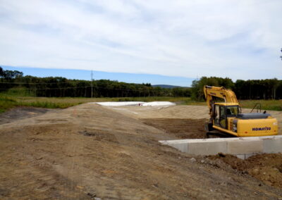 Graded dirt construction site with long, flat-bottomed depression. A horizontal concrete berm is in the foreground and a backhoe.