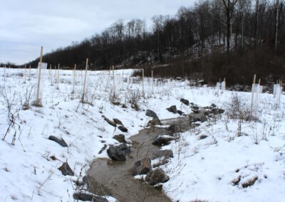 Muddy water flowing in winding section of stream channel with banks covered by snow.