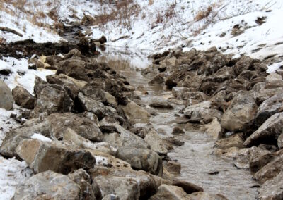 Muddy water flowing amid rock-lined stream channel in winter.