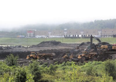 Landscape view of several pieces of heavy equipment (dumptruck, excavator, bulldozer) working on dirt construction site with uneven topography. A warehouse in operation is in the background.