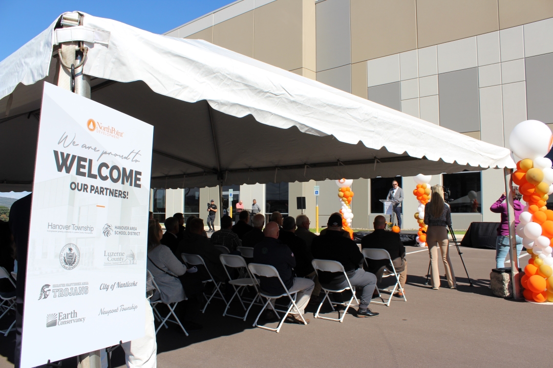 Under a white tent set up in a parking lot, a group of seated individuals listens to a man speaking at a podium. A new, large warehouse is behind him. Several columns of orange and white balloons decorate the area.