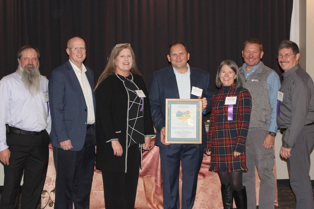 Five adult men and two women stand in a line posing for picture. Man in middle (Terry Ostrowski) holds a framed award document.