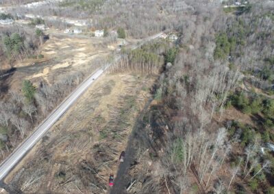Aerial view of road passing through a partially cleared forest within the Nanticoke Creek Watershed; logs and tree debris are scattered on the ground with vehicles visible, surrounded by remaining woods and some houses in the distance.