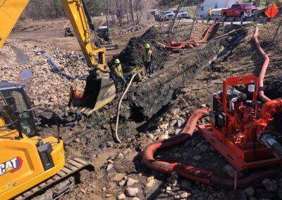 Several construction workers stand by a large trench. An excavator is digging, and red and yellow hoses are laid around the site. Equipment and rocks surround the work area.