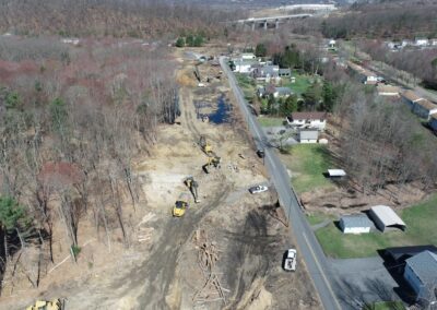 Aerial view of a construction site beside a road, with several excavators working on cleared land within the Nanticoke Creek watershed. Nearby are houses, trees, and vehicles parked along the street.