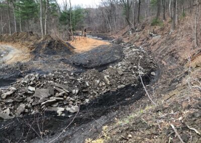 A roughly graded trench runs alongside large pile of rocks and soil in a wooded landscape with bare trees in the background. The scene suggests recent earth-moving or construction work.