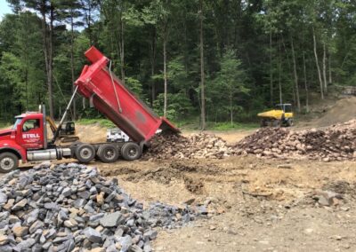 A red dump truck unloads a large pile of rocks onto the ground at a construction site surrounded by trees.
