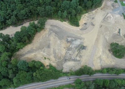Aerial view of a large cleared and leveled construction site bordered by trees, with a paved road in the foreground. The surrounding area is mostly wooded.