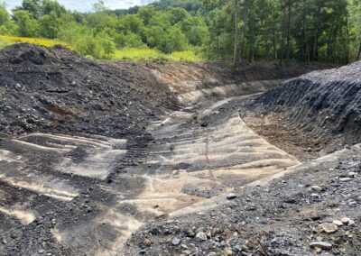 A large, winding concrete flume with tall piles of rock and soil an either side. Trees and dense greenery visible in the background.