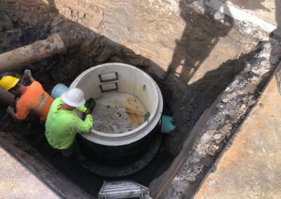 Two workers in safety gear install a large concrete structure in a deep trench within t. A metal ladder leads into the trench and pipes are visible.