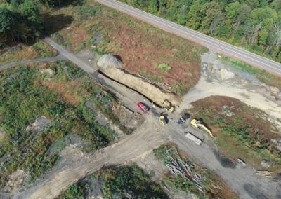 Aerial view of a construction site within the Nanticoke Creek watershed, featuring excavators, trucks, a long trench, and cleared land near a road. The surrounding area is partly forested, with scattered logs and construction equipment visible.