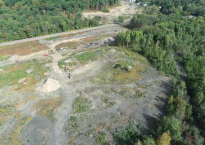 Aerial view of a construction site with several vehicles and equipment on a cleared, dirt-filled area. Trees and a road border the site.