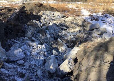A large dirt area filled with broken concrete and debris. Patches of snow and dry grass are visible in the background