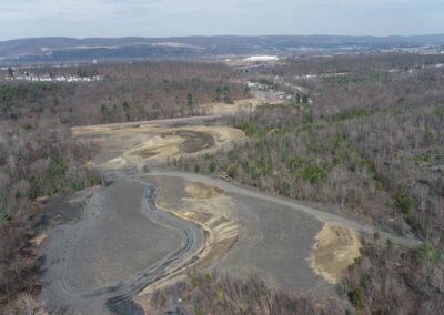 Aerial view of a cleared construction area with long, serpentine channel in the middle. The site is by forested land. Roads and a few buildings are visible in the background.