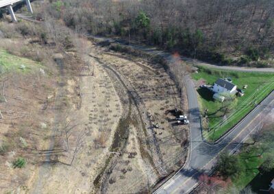Aerial view of a narrow, creek bed next to a road within the Nanticoke Creek watershed; a white house sits nearby, and a few vehicles and equipment are present along the road. The surrounding area is wooded.