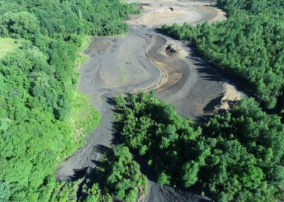 Aerial view of a deforested area with exposed soil graded into a serpentine trench, surrounded by dense green forest. Construction vehicles are visible in the distance on the cleared ground.