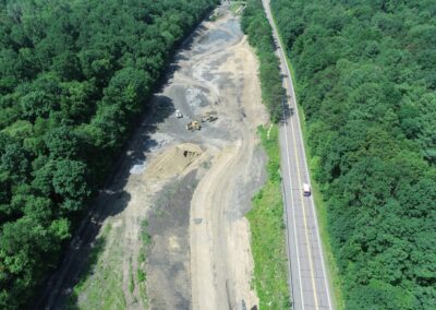Aerial view of a construction site with earth-moving equipment working on grading a long, winding channel within the Nanticoke Creek watershed. The site is bordered by forest and a two-lane road.