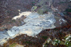 Aerial view of small, forested mountainside in autumn. At center, a large portion is barren and unnaturally shaped, with a pit in the lower left-hand corner. Construction vehicles can be seen.