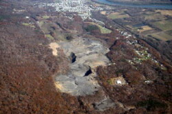 Aerial view of large barren dirt site with two massive pits in it. Surrounded by rust-colored forested areas. A town alongside a river is at upper edge.