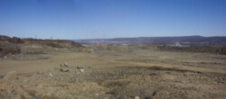 Landscape view of large, flat, dirt area with some piles of rocks to side. Site overlooks valley and small towns can be seen in distance.