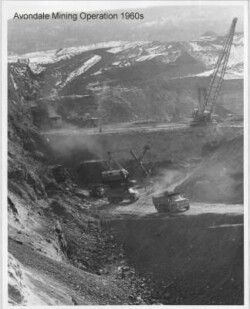 Black and white landscape view of strip mining, consisting of crane, excavators, and trucks on tiers carved into side of pit