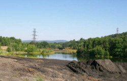 Landscape view of pond surrounded by green forested area. Land in front is mainly flattened dirt, although a large pile of silt remains to right.