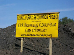 Large yellow sign posted in dirt. Wording reads: "Franklin Bank Reclamation Project / Work funded by EPA Brownfields Cleanup Grant / and Earth Conservancy"