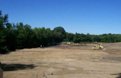 Heavy construction vehicles move across large dirt site, lined by green trees.