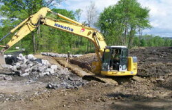 Excavator (center), working in dirt area, moves large rocks piled on left-hand side of image.
