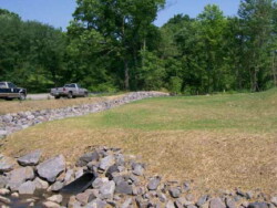 Grassy area lined in the back by large deciduous trees. A rock lined channel runs along left-hand side. Two pick-up trucks are next to it.