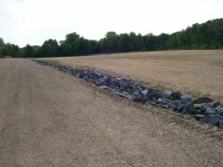 Landscape view of flat, recently seeded, brown land, bisected by a channel filled with larger-sized rocks. Back of site is tree-lined.