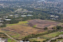 Aerial of large, cleared, flat piece of land. Roadways run along two sides of property. Warehouses and residential areas surround site.