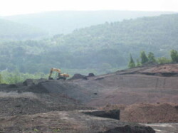 Landscape view of barren, uneven land with excavator working in distance against the background of a rolling green, forested mountainside