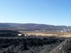 Landscape view of several construction vehicles and pickup trucks on flattened dirt with many piles of waste rock and silt in front and behind them