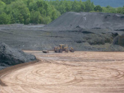 Heavy construction equipment (backhoe with roller) flattens dirt area, marked with circular track marks. Large banks of waste coal tower above it in the background.
