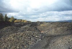 Landscape view of rocky, undulating piles of mine spoils. A forested area is in the background on the left-hand side. A sheet of clouds covers the sky.
