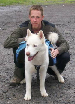 A man crouches down behind a white shepherd dog outdoors on gravel area.