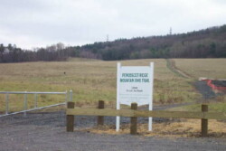 Image of white sign with green lettering indicating trailhead for Penobscot Ridge Mountain Bike Trail. A path runs behind the sign through a field at a slight incline towards a forested area.