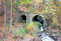 Small double-arched bridge, under hill, made of concrete and stones in woods. Autumn-colored leaves cover the ground. Creek runs through right arch of bridge.