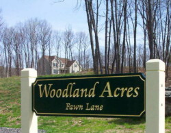 Green sign with gold calligraphic writing hanging between two white posts. Sign reads: "Woodland Acres / Fawn Lane." A new, two-story house can be seen in the background.