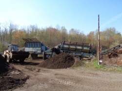 Large blue pieces of composting equipment. One reads "power screen trommel." A loader is to their left. A pile of dirt-like material (compost) is in front of them.