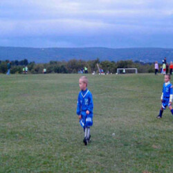 Several elementary-aged boys in royal blue uniforms playing soccer on a field. A net, adults are in background.