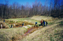 Landscape view of winding orange creek surrounded by grass with several college students scattered around it. Some are bending/looking toward water.