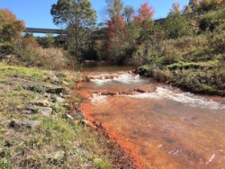 Landscape view of creek going over 2-3 steps of rocks. Rocks, stream bed, and immediate bank are orange (acid mine drainage).